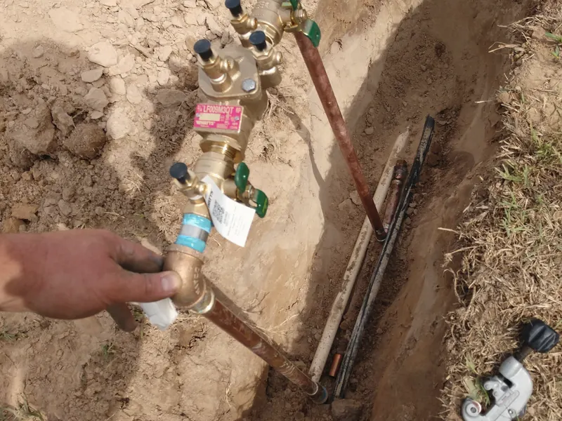 A hand holds a new brass backflow assembly on a water line in an open trench.