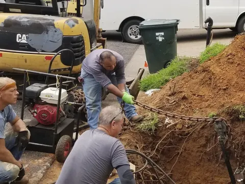 Four men in work clothes dig out a pipe trench in a front yard; a white box van and a small excavator stand behind them.