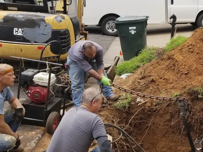 Four men in work clothes dig out a pipe trench in a front yard; a white box van and a small excavator stand behind them.