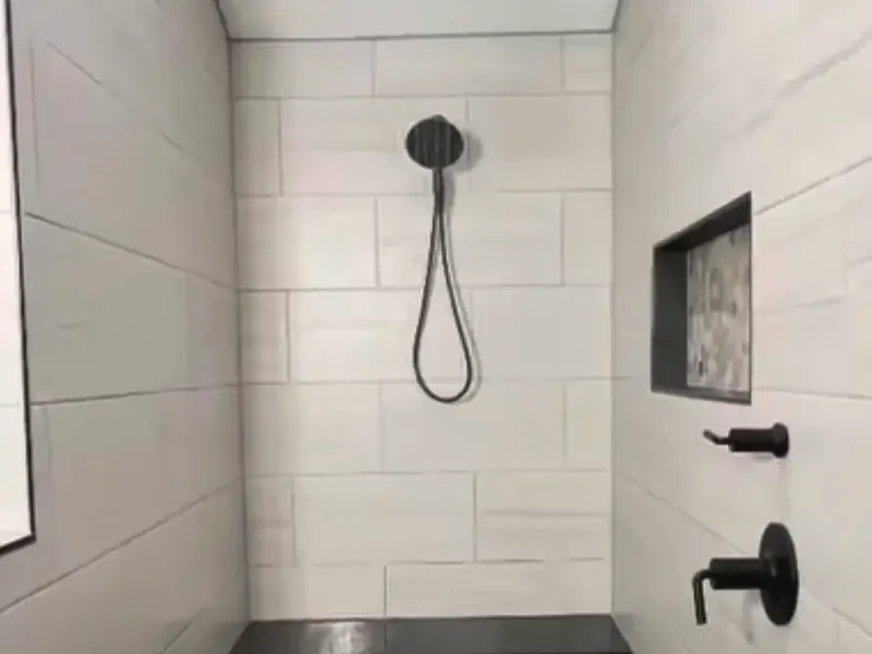A finished walk-in shower with white tile, a black rain head, a tiled bench and a pebble floor.