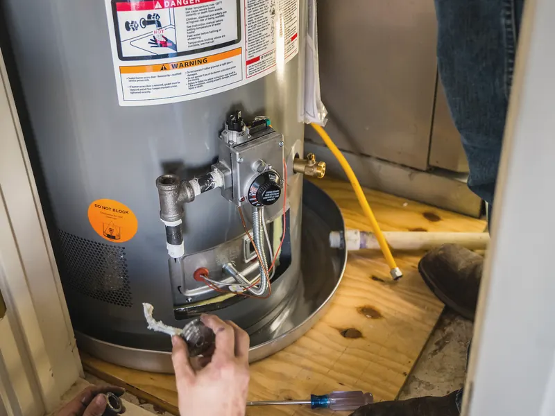 A technician works on the gas valve at the base of a water heater, seen from above.