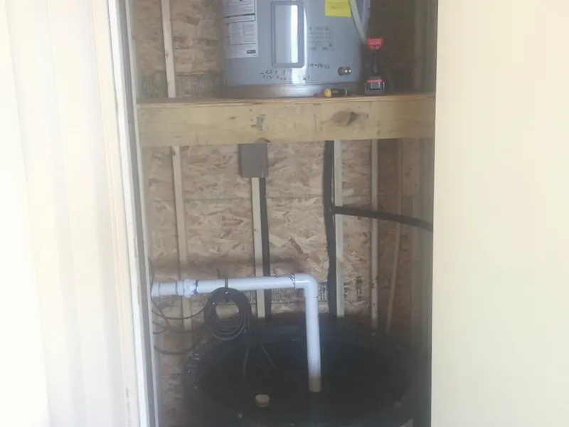 A water heater on a shelf above a sump tank with new PVC piping, inside a small utility closet.