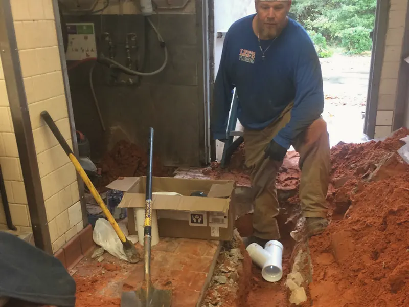 A plumber kneels in a trench dug through a building floor to reach a sewer line.