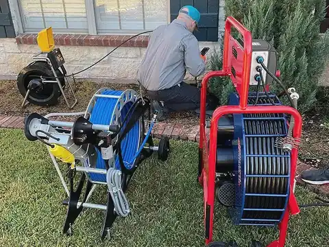 A technician kneels by a house with drain-cleaning and camera equipment on the lawn.
